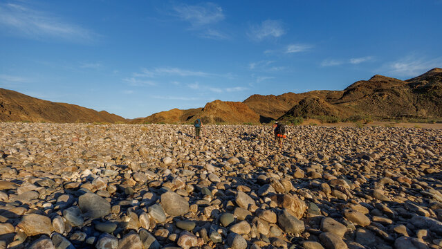 View of hikers trekking across the rocky riverbed of Fish River Canyon with arid mountains in the background under a clear blue sky in Hobas, Karas Region, Namibia.