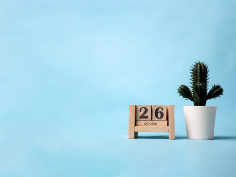 A wooden block calendar for October 26th with a plastic cactus plant on the side on a light blue background.