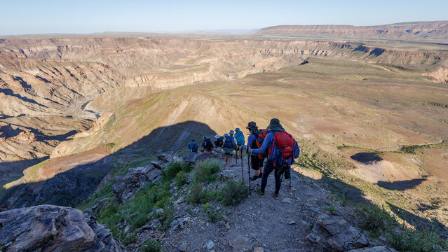 View of a group of hikers with backpacks and trekking poles walking along a rocky trail overlooking the vast Fish River Canyon in Hobas, Karas Region, Namibia.