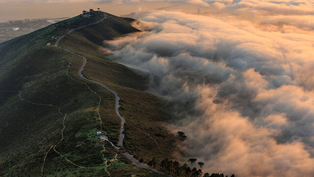 View of the winding road leading up Lion's Head mountain during golden hour with thick clouds rolling over the slopes in Cape Town, Western Cape, South Africa.