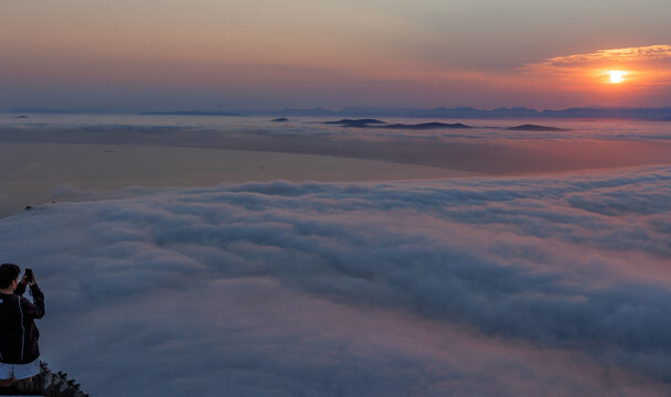 View of a person taking a photo with a smartphone from the summit of Lion's Head overlooking a vast sea of clouds during a golden sunset in Cape Town, Western Cape, South Africa.
