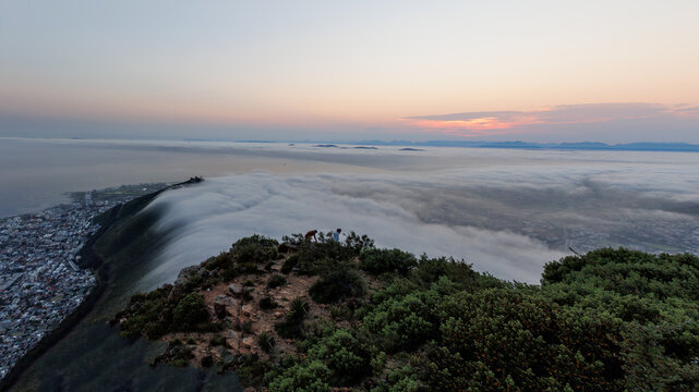 View of Lion's Head in Cape Town with a thick layer of clouds rolling over the mountain at sunset, overlooking the city lights in Cape Town, Western Cape, South Africa.