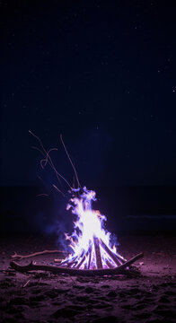 Campfire burning on sandy beach at night with glowing violet flames and embers under dark night sky