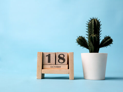 A wooden block calendar for October 18th with a plastic cactus plant on the side on a light blue background.