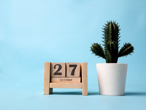 A wooden block calendar for October 27th with a plastic cactus plant on the side on a light blue background.