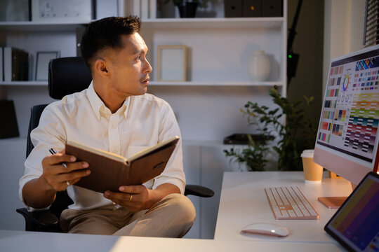 Asian male designer reviewing color samples and documents at his office desk.