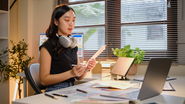 Asian female designer reviewing color documents while working with a laptop and tablet.