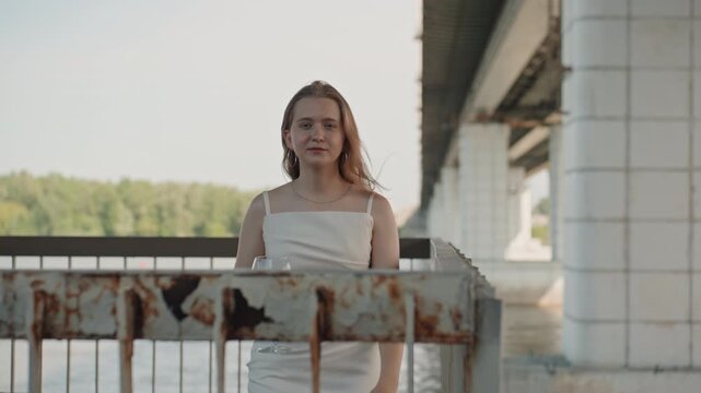 Woman in white dress stands on industrial bridge holding and raising glass of red wine. background features water greenery and underside of large concrete pillars under soft daylight