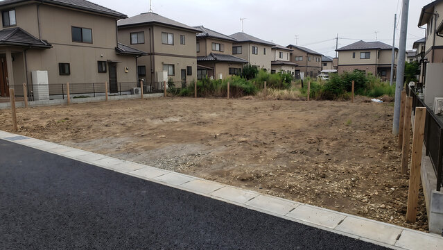 舗装道路に接する広い砂質土の住宅街空き地と周辺戸建て住宅、木製杭と区画境界の景観