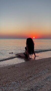 A brunette woman with long, disheveled hair, wearing a black two‑piece swimsuit, is sitting on the sand by the sea at sunset; the view is from behind.