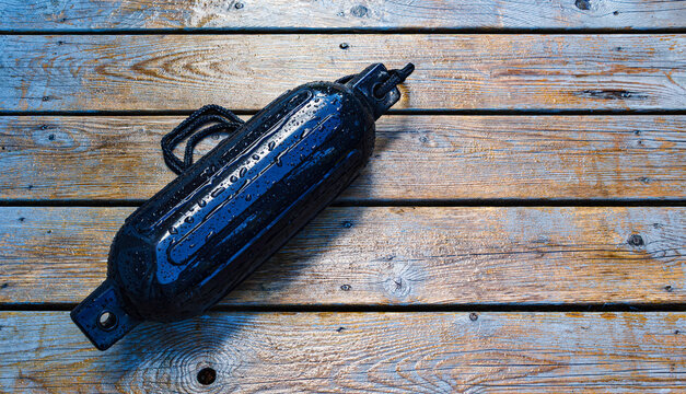 Black cylindrical boat fender rests on a weathered and wet wooden dock.