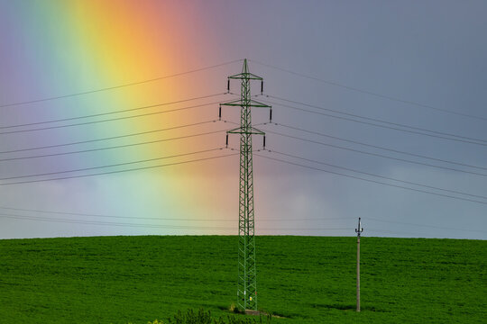 View of an electricity pylon standing in a lush green agricultural field under a cloudy sky with a vibrant rainbow in the background in Brno, South Moravian Region, Czechia.
