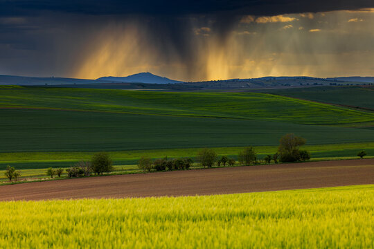 View of rolling agricultural fields under dramatic storm clouds with golden sunlight illuminating the distant hills in Brno, South Moravian Region, Czechia.