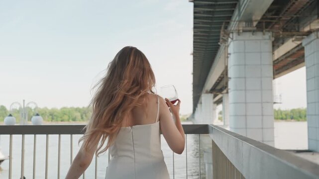 Woman in white dress holds and sips red wine while standing on bridge with water greenery and underpass visible. Her hair moves naturally as woman looks around creating relaxed atmosphere