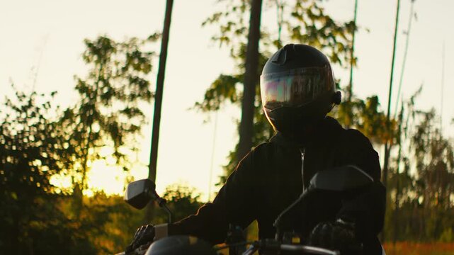 Motorcyclist in black gear riding through golden countryside at sunset, reflective helmet visor glowing in warm evening sunlight with cinematic lens flares
