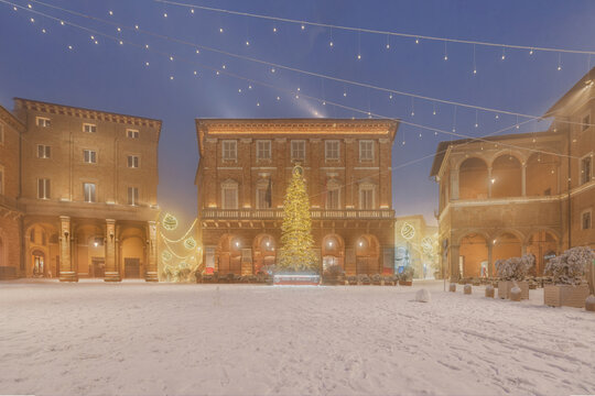 View of a large illuminated Christmas tree in the snow-covered Piazza della Liberta, surrounded by historic buildings with porticoes and festive string lights, Macerata, Marche, Italy.