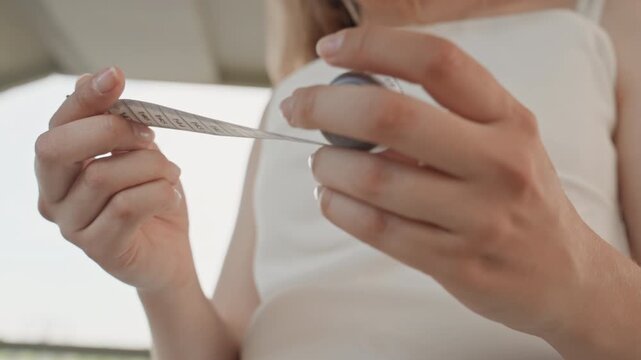 Woman in white dress measures her body with tape measure focusing on hands and torso. scene is softly lit indoors or near natural light emphasizing texture detail as woman unrolls