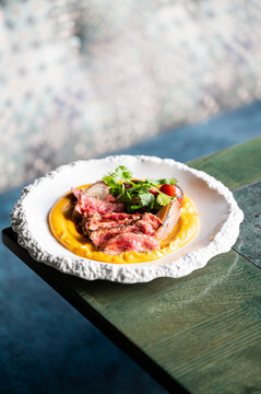 Sliced medium-rare beef steak on a bed of yellow puree garnish with fresh herbs on a white ceramic plate