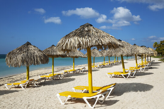 View of yellow lounge chairs and straw umbrellas on a white sand beach with turquoise water under a blue sky at the Playa Ancon beach in Trinidad, Sancti Spiritus, Cuba.