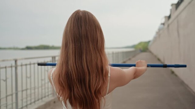 Woman with long brown hair walks along riverside path holding blue pole of unattended rowing boat. Woman wears white sleeveless top and moves steadily forward on concrete beside metal