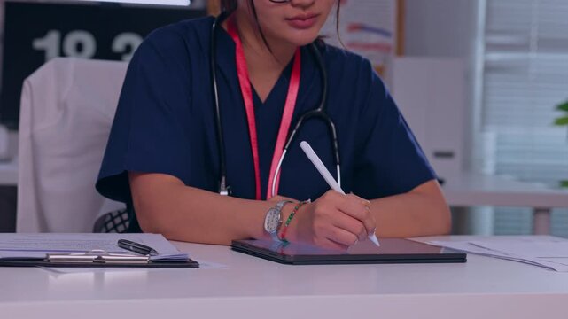 Medical Professional Writing Patient Notes at Desk with Stethoscope, Navy Uniform, Red Lanyard, Tablet, and Identification Badge in Modern Healthcare Clinic Office