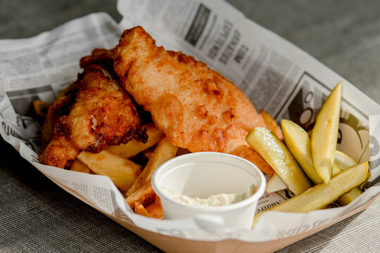 Plate of fish and chips, the famous British classic consisting of fish fillets in golden batter and french fries, served with a small bowl of sauce on a sheet of paper resembling newspaper print