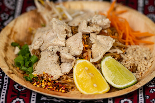 Chicken Pad Thai, a popular Thai stir-fried rice noodle dish, garnished with sliced ​​chicken, lime, crushed peanuts, and scallions, served in a bamboo dish
