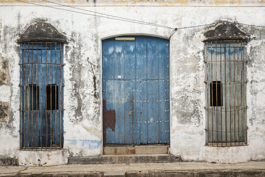 View of a weathered white wall featuring a central blue wooden door flanked by two blue windows with metal grilles in Remedios, Villa Clara, Cuba.
