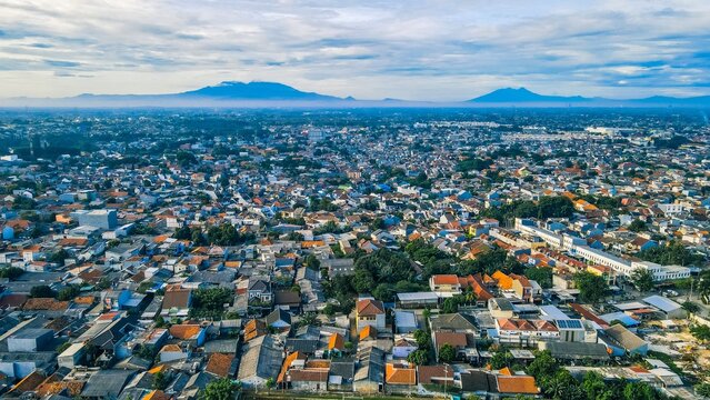 Aerial view of the dense urban sprawl featuring residential rooftops, distant mountains, and a clear sky in Bekasi Barat, West Java, Indonesia.