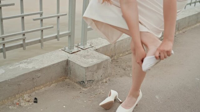 Woman removes white high heeled shoes and puts on socks by riverside walkway. Woman lifts her dress to tuck in sock with casual sneakers nearby on concrete pavement near metal railings