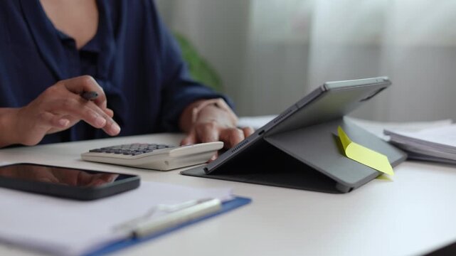 Asian businesswoman sitting at desk using digital tablet with stylus while analyzing financial charts and typing on laptop computer in bright modern office space room environment