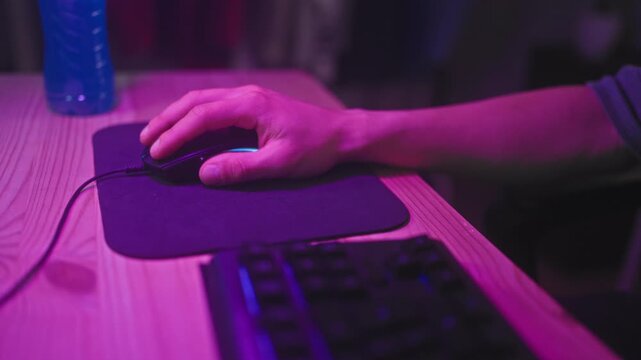 Man holding a wired mouse with right hand on a mousepad at a wooden desk beside a keyboard and bottle in a purple lit studio; concentration.