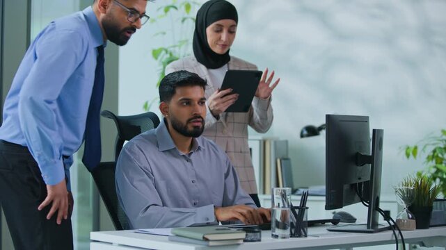 Arabian leader mentor boss with secretary coming helping to intern man with business work looking in computer talking advice showing thumb up three colleagues team group discuss online task in office