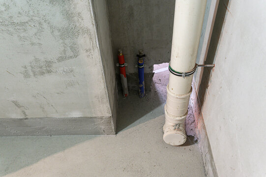 Beige PVC drain pipe with couplings and wall clamp, red and blue supply lines in recess, rough concrete walls, raw utility corner, industrial plumbing layout