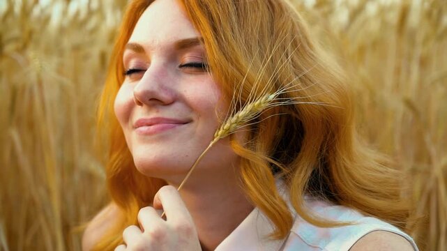 Dreamy young ginger woman with closed eyes holding golden ear of wheat near cheek. Smiling female model relaxing in summer field scenery