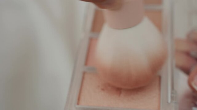 Hand holding makeup brush repeatedly sweeps across different shades of pink and peach blush compacts in open palette demonstrating application process. close up view highlights soft lighting