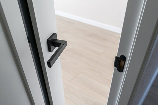 Matte black geometric door handle on white door with black trim, visible latch plate, clean angular lines, minimalist design, light wood floor, neutral interior space