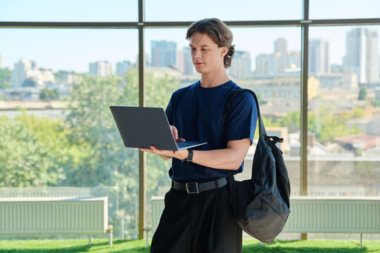 Young male student standing with backpack and working on laptop near window