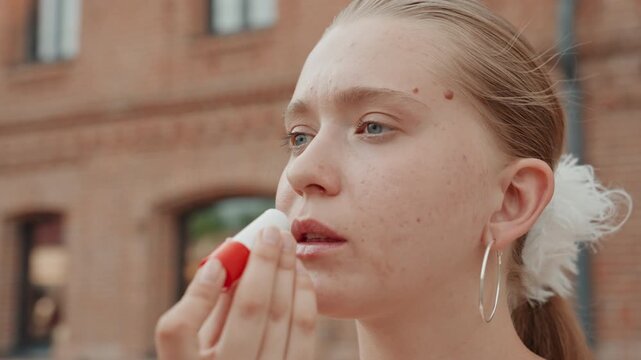 Young woman applies red lip balm outdoors in front of brick building with arched windows. Woman has light brown hair tied back wears large hoop earrings and soft natural makeup