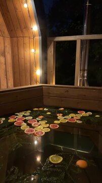 A wooden hot tub on the porch of a country house, surrounded by garlands filled with citrus, fir branches in the evening. Enjoying a moments of relaxation in the open-air bath