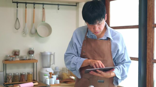 Young Asian Man in Apron Checking Cookie Recipe on Tablet in Home Kitchen