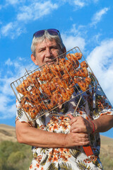 Man holds skewers of grilled meat outdoors. Sky blue and bright, clouds drift above. He wears colorful shirt and sunglasses. Sunlight glows warmly on his smiling face. Perfect for food, travel © kvdkz