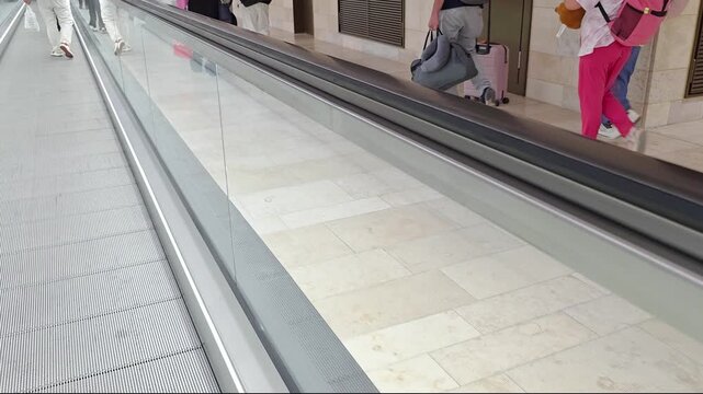 Passengers with suitcases walk beside a moving walkway through the airport arrivals area, travel transit scene, airport terminal movement.