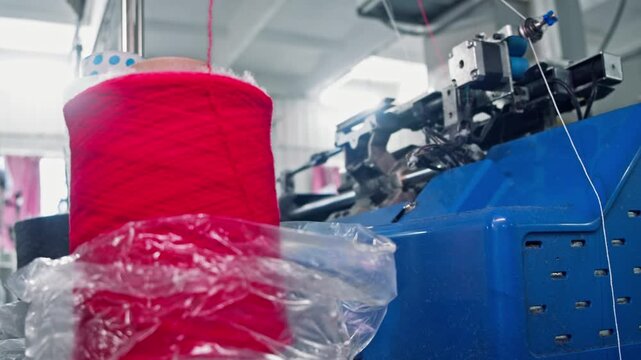 Close up of red yarn spool feeding thread into industrial textile machine during automated production. Moving mechanical parts process yarn inside factory workshop.
