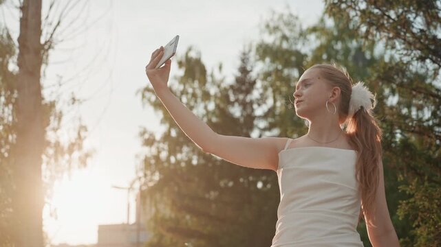 Young woman takes selfie outdoors during sunset adjusting her hair while holding phone. Woman wears white dress and large hoop earrings with sunlight backlighting through trees in soft focus