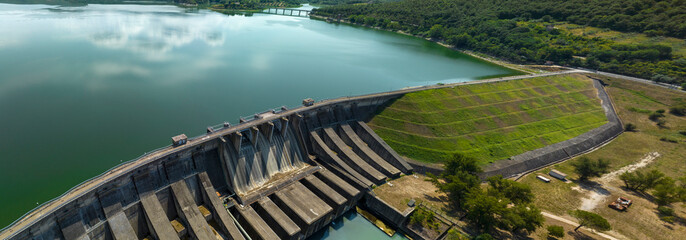 Obraz premium Aerial view of a large concrete dam with spillway gates, a grassy embankment, and a bridge crossing a calm reservoir, surrounded by forested hills. It's Corbara Lake located in Umbria, Italy.
