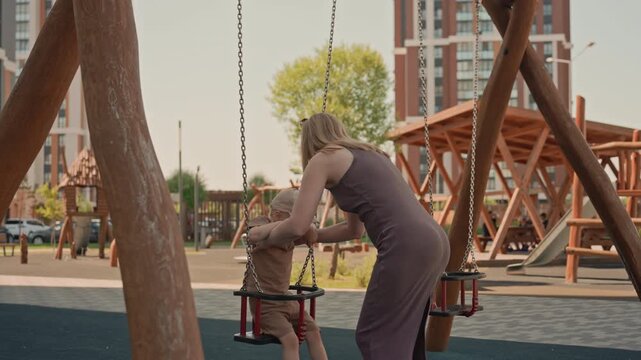 Woman gently pushes young boy on swing at outdoor playground. child holds onto chains while wearing casual clothing and both are surrounded by wooden play structures under daylight