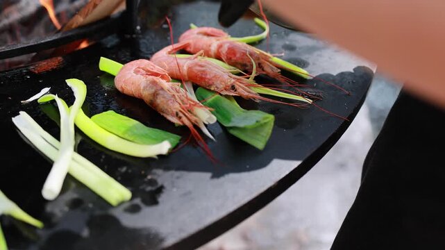 Shrimp and leek grilling on round fire plate. Fresh prawns cooked over open flame outdoors. Seafood barbecue preparation with vegetables on hot steel surface
