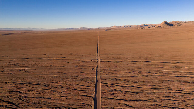 Aerial view of a long straight road stretching through the vast arid landscape of the Atacama desert under a clear blue sky in Chile.