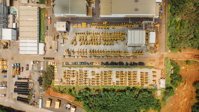 Aerial view of a large manufacturing facility with rows of yellow construction cranes and heavy machinery parked in an organized yard, surrounded by industrial buildings and green trees in India.